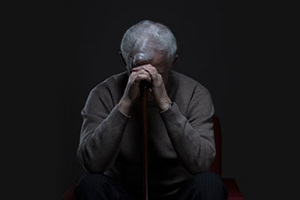 An elderly man sitting in a chair, resting his hands on his face, appears deep in thought